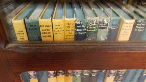 A close-up of vintage "Tom Swift" book spines displayed in a wooden bookshelf.