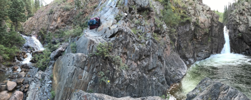 A panoramic view of rocky cliffs with two waterfalls cascading into a serene pool surrounded by lush greenery.
