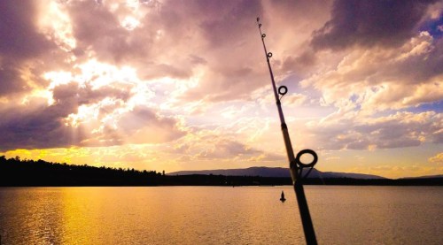 A fishing rod points towards a colorful sunset over a calm lake, with clouds and distant mountains in the background.