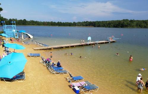 A sunny beach scene with people swimming, lounging on chairs, and a slide near a dock by a calm lake.
