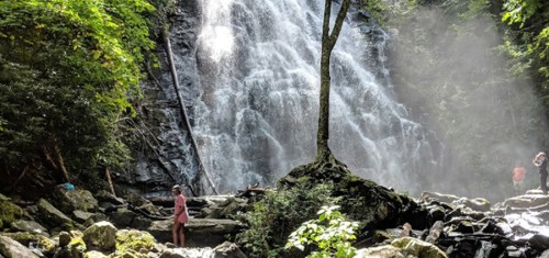 A waterfall cascades down rocky cliffs, with two people exploring the misty area surrounded by lush greenery.