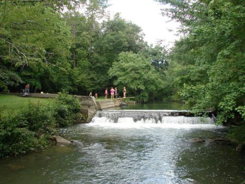 A serene river scene with a small waterfall, surrounded by lush greenery and people enjoying the outdoors.