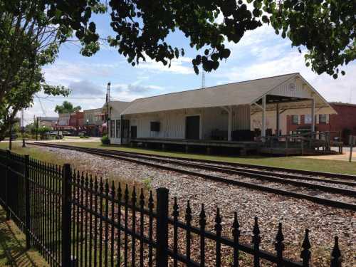 A train station building beside railway tracks, surrounded by greenery and a black fence under a clear blue sky.