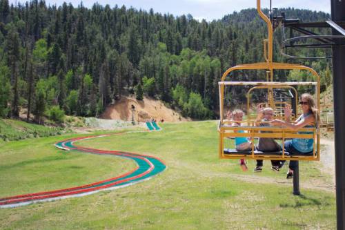 A family rides a yellow chairlift over a green landscape with a winding track and trees in the background.