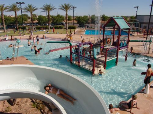 A busy water park with a slide, pool, and children playing in the water under sunny skies and palm trees.