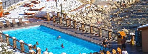A snowy landscape surrounds a pool with people swimming, alongside a rocky riverbank and lounge chairs.