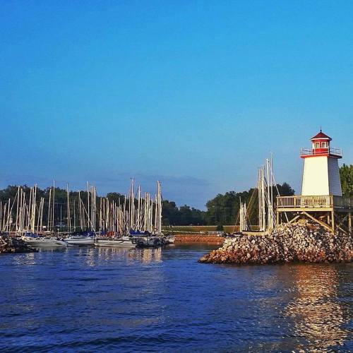 A lighthouse stands on a rocky shore, with sailboats docked in the harbor and trees in the background under a clear blue sky.