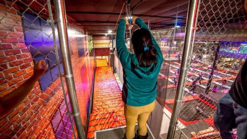 A person prepares to zip line over a colorful indoor trampoline park, surrounded by vibrant walls and safety fencing.