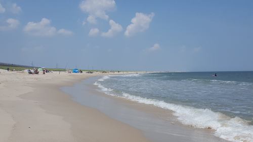 A sunny beach scene with gentle waves, sandy shore, and a few people enjoying the day under a clear blue sky.