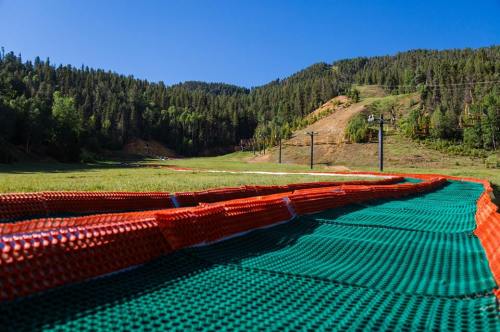 A green and orange ski slope with a chairlift in the background, surrounded by lush forested mountains under a clear blue sky.