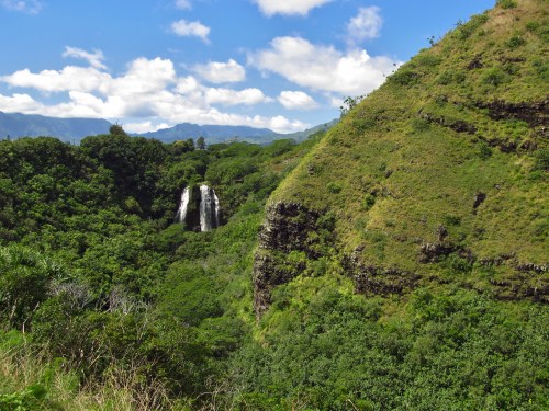 A lush green landscape featuring a waterfall cascading down a rocky cliff, surrounded by hills and blue skies.