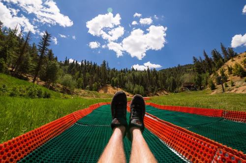 A person's legs on a green and orange slide, with a forested hillside and blue sky in the background.
