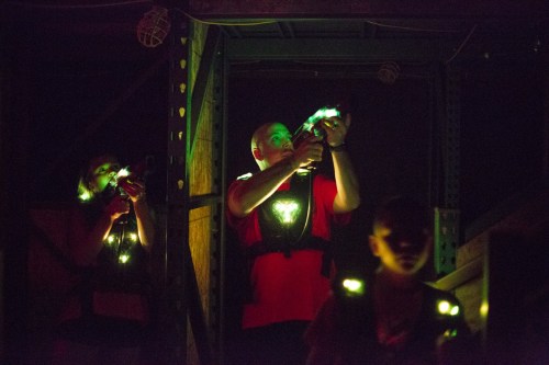 Two adults and a child in dark vests aim laser guns in a dimly lit indoor arena. Green lights illuminate their faces.