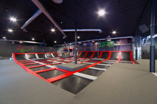 A spacious indoor trampoline park with red and gray trampolines, surrounded by black walls and bright overhead lights.