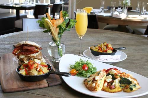 A table setting with a sandwich, two egg dishes, roasted vegetables, a salad, and a mimosa in a restaurant.