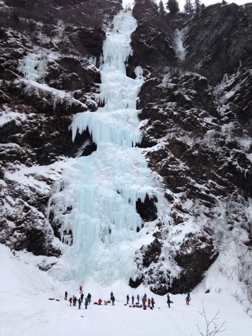A frozen waterfall cascades down a rocky cliff, with a group of people gathered at the base in a snowy landscape.