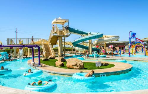 A vibrant water park scene with slides, a lazy river, and children playing in the pool under a clear blue sky.