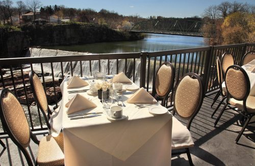 A scenic restaurant balcony overlooking a waterfall and bridge, set with a table for dining.