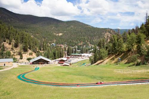A scenic view of a hillside amusement park with a winding track, surrounded by trees and mountains under a blue sky.