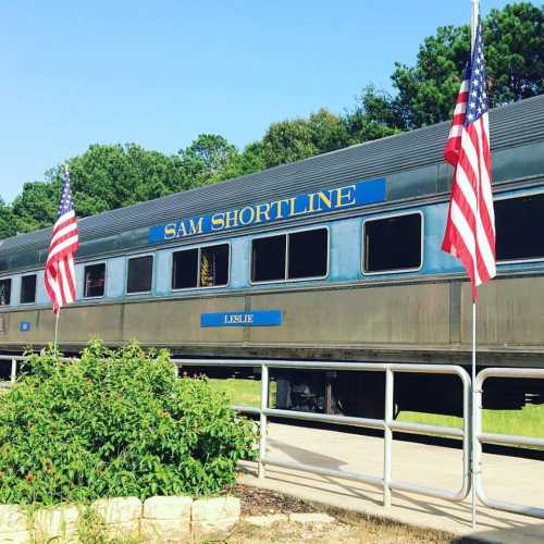 A vintage train car labeled "SAM SHORTLINE" with American flags on either side, surrounded by greenery.