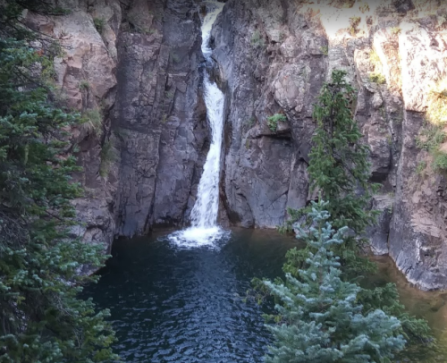 A waterfall cascades between rocky cliffs into a serene pool, surrounded by lush green trees.