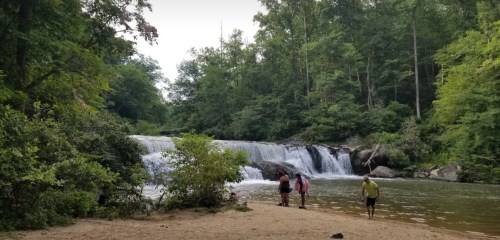 A serene waterfall surrounded by lush greenery, with people enjoying the water and sandy shore.