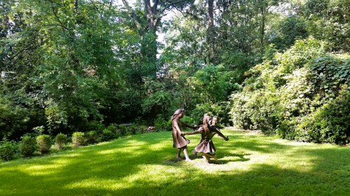 Two bronze statues of children playing in a lush green garden surrounded by trees and shrubs.