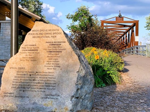 A stone monument with engraved text, flowers, and a wooden bridge in the background at Inver Grove Heights Recreational Pier.