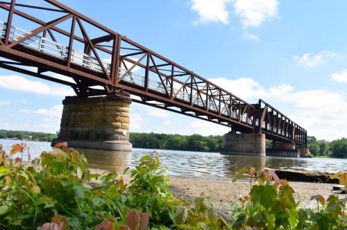 A rusted metal bridge spans a river, supported by stone pillars, with greenery in the foreground and blue skies above.