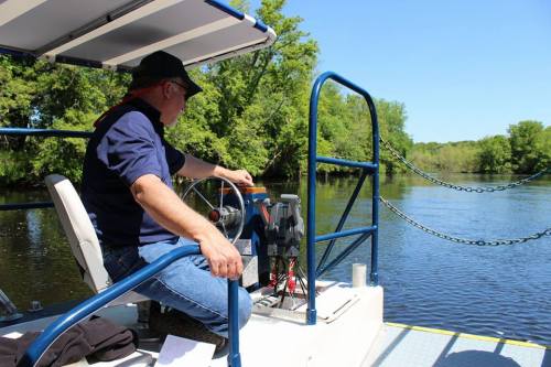 A person sits at the helm of a boat on a calm river, surrounded by lush greenery and clear blue skies.