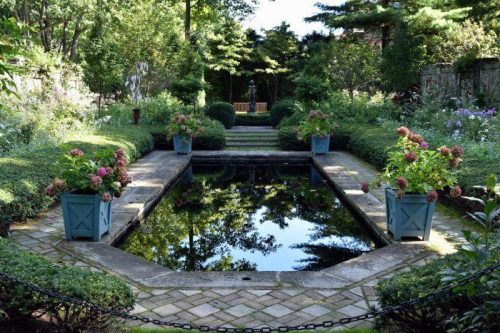 A serene garden with a reflecting pool, surrounded by lush greenery and flower-filled planters under a clear blue sky.