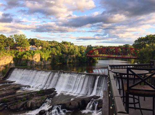 A scenic view of a waterfall with a red bridge in the background, surrounded by lush greenery and a cloudy sky.