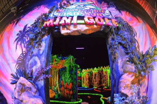 Entrance to a vibrant, neon-lit mini-golf course themed around dinosaurs, surrounded by colorful decorations.