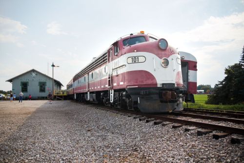 A vintage train in maroon and white parked beside a historic train station on a sunny day.