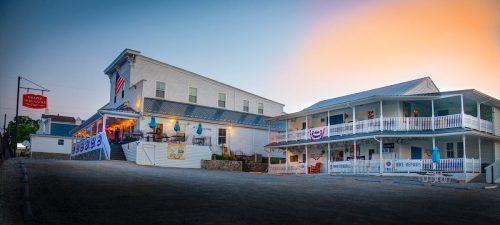 A charming building with a porch, flags, and outdoor seating, set against a colorful sunset sky.