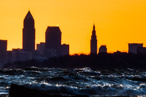 Silhouette of a city skyline against a vibrant orange sunset, with waves in the foreground.