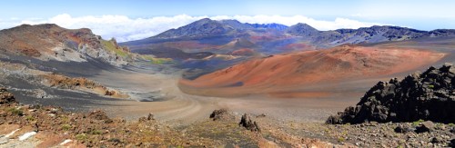 A panoramic view of a volcanic landscape with colorful slopes, rocky terrain, and distant mountains under a blue sky.