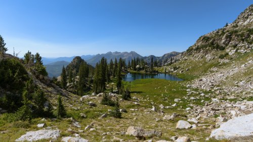 A serene mountain landscape featuring a clear lake surrounded by rocky terrain and lush green trees under a blue sky.