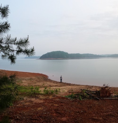 A serene lakeside view with a person standing on the shore, surrounded by trees and distant hills.