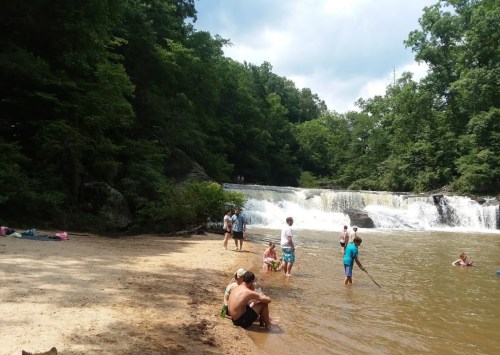 A sandy beach by a waterfall, with people swimming and playing in the water surrounded by lush green trees.