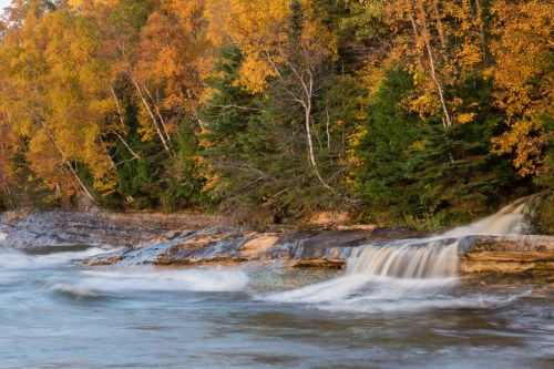 A serene river flows over rocks, surrounded by vibrant autumn foliage in shades of orange and green.