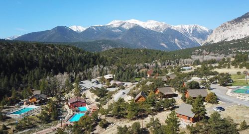 Aerial view of a mountain resort with cabins, pools, and snow-capped peaks in the background. Clear blue sky above.