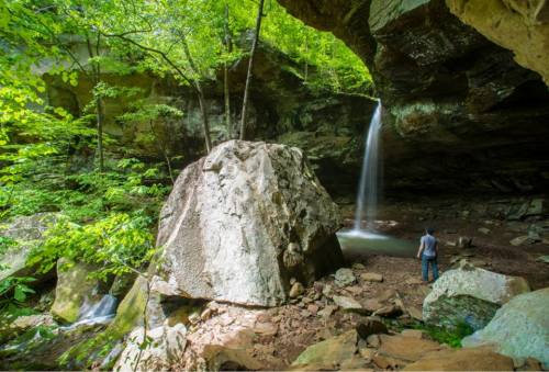 A person stands near a waterfall cascading from a rocky overhang, surrounded by lush green trees and boulders.
