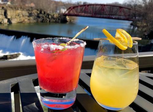 Two colorful cocktails sit on a table with a scenic river and bridge in the background on a sunny day.