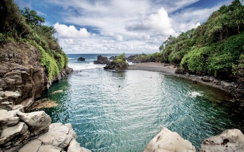A serene coastal scene with clear water, rocky formations, and lush greenery under a partly cloudy sky.