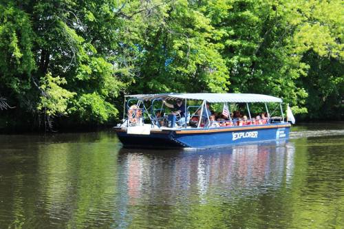 A tour boat named "Explorer" glides through a calm river surrounded by lush green trees.