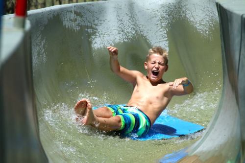 A young boy slides down a water slide, splashing into the water with an excited expression.