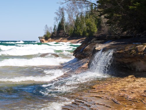 A rocky shoreline with gentle waves, a small waterfall, and lush trees under a clear blue sky.