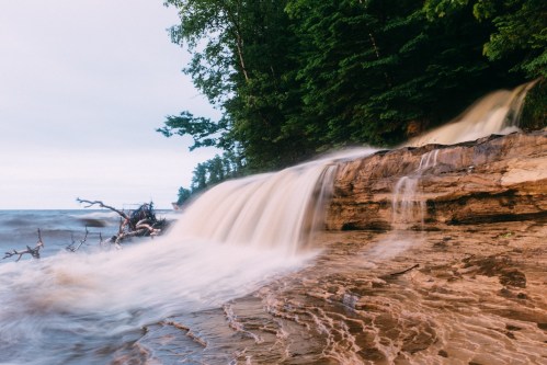 A serene waterfall cascades over rocky terrain into a calm lake, surrounded by lush green trees.