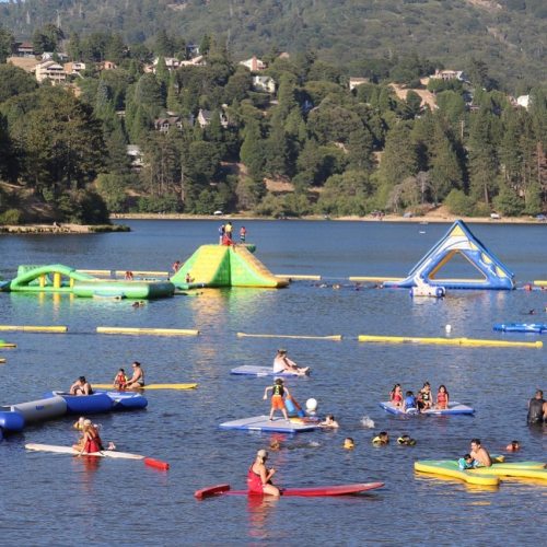 A sunny lake scene with people swimming, paddleboarding, and enjoying inflatable water attractions. Trees and hills in the background.
